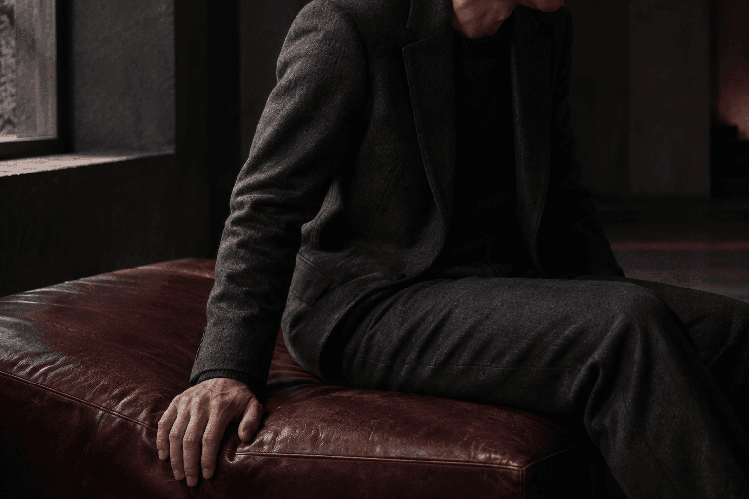 A seated figure in charcoal wool, one hand resting across worn oxblood leather beneath a compressed seat cushion.