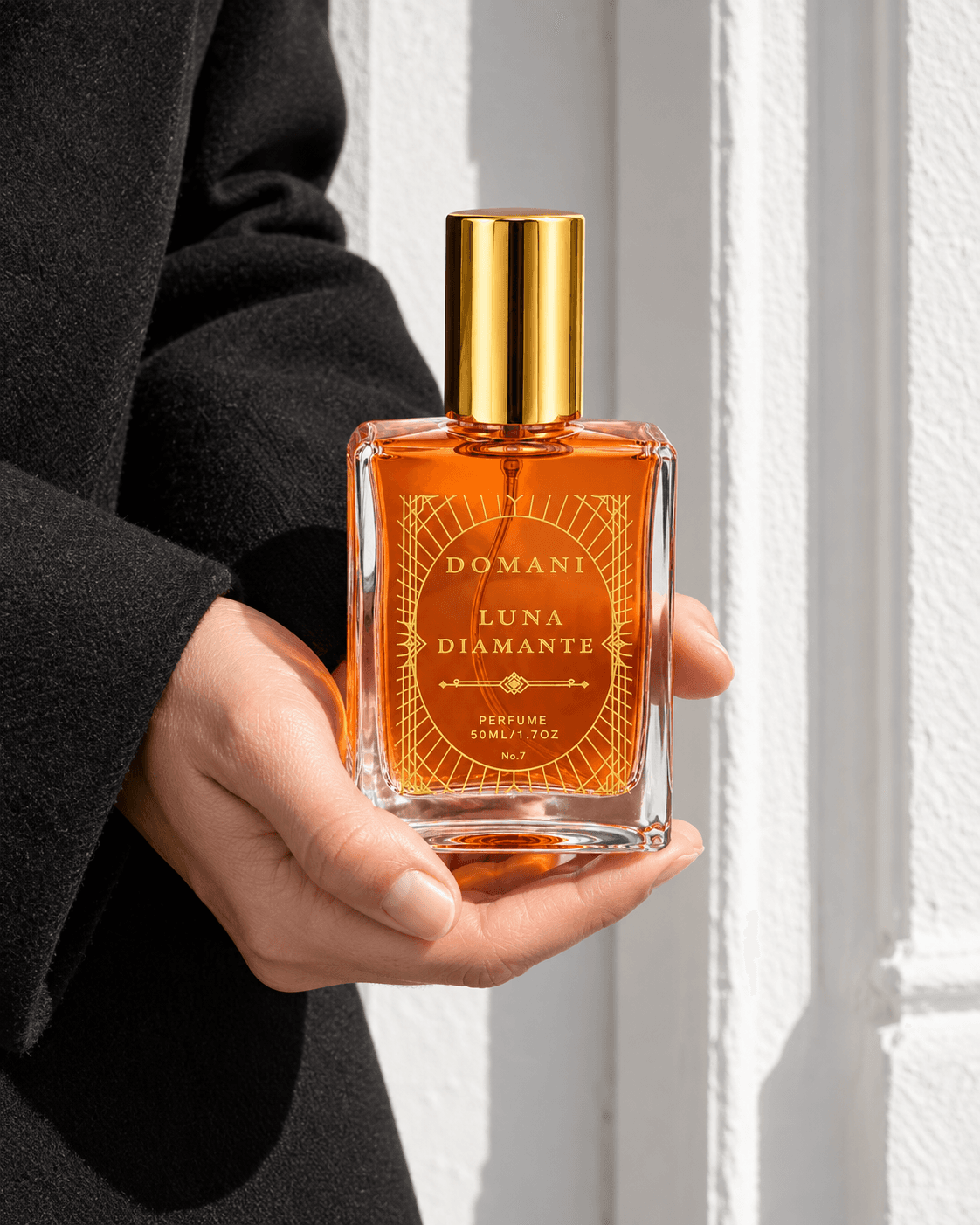 A hand supports the complete Luna Diamante 50 ml perfume bottle, with orange liquid and fine gold artwork against black wool and a white doorway.