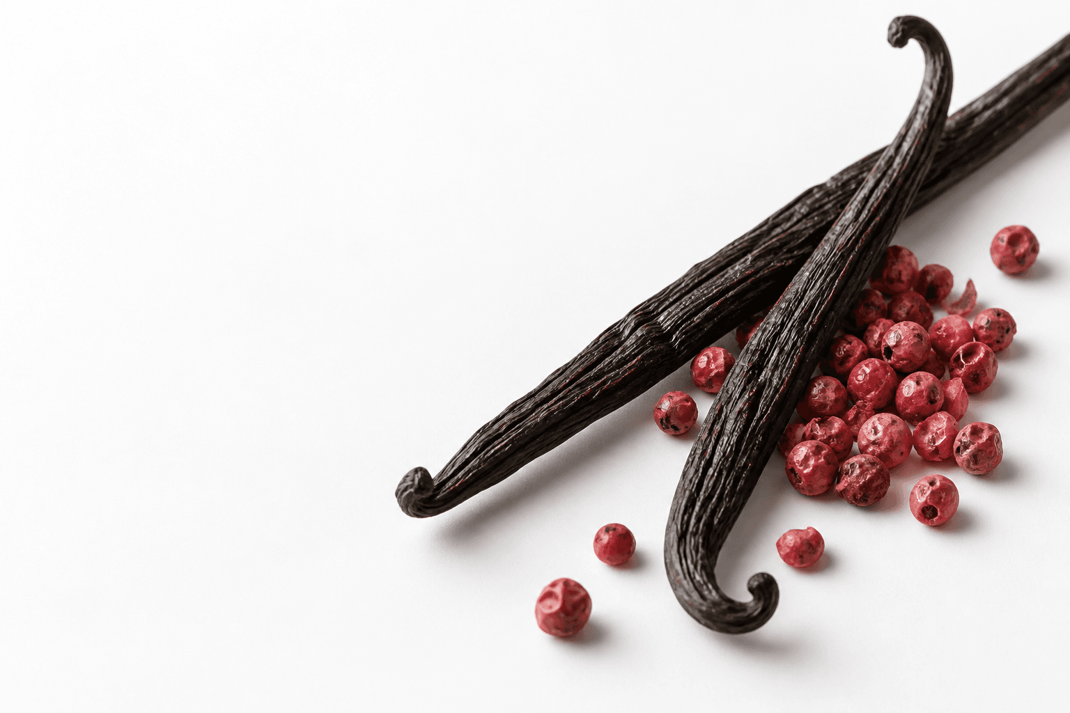 A close view of wrinkled vanilla bean pods and pink peppercorns on a white photographic surface.