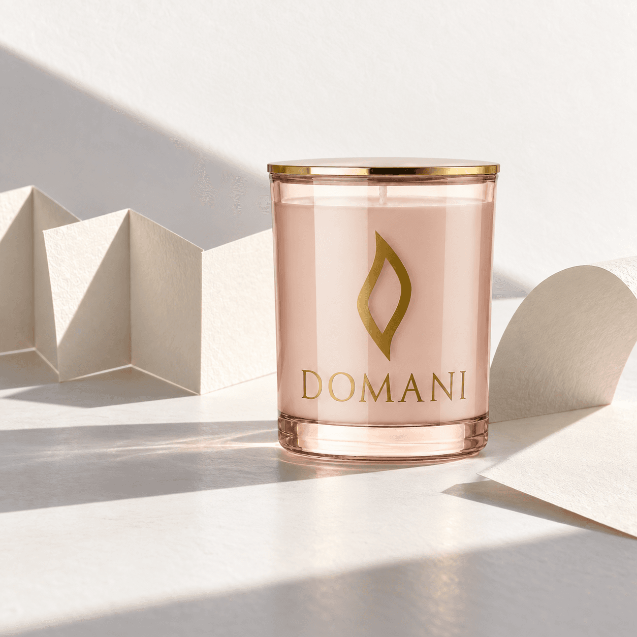 The complete unlit Novella candle in Rose Quartz glass with its gold lid on, beside folded ivory paper and a curved sheet casting crisp shadows on white.