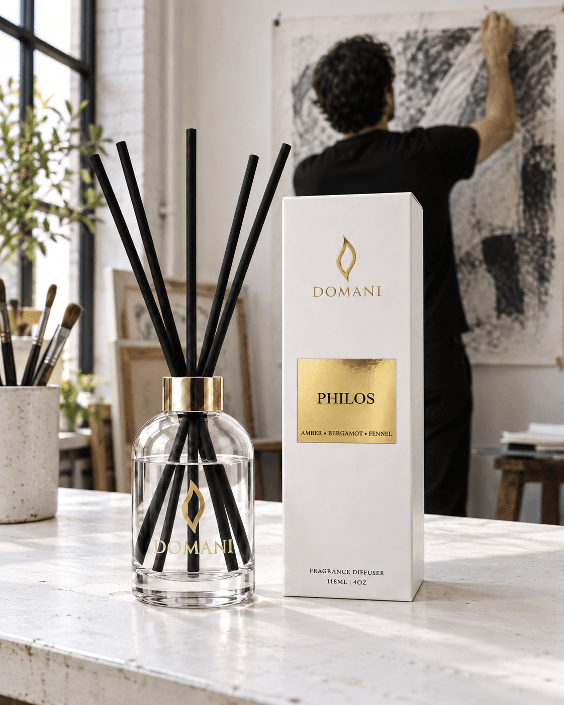 Philos reed diffuser and its named white and gold carton on a studio workbench, with an adult hanging a monochrome paper study beyond.
