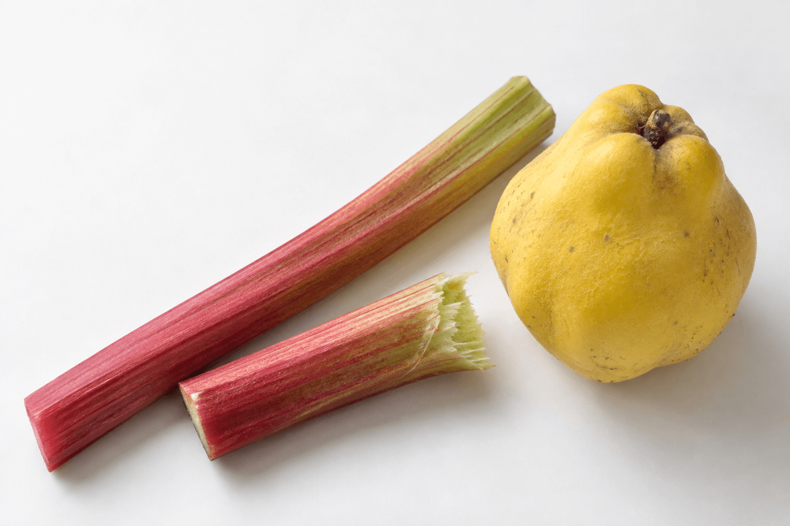 A yellow quince beside red and green rhubarb stalks, with a torn end exposing pale fibers on a white surface.