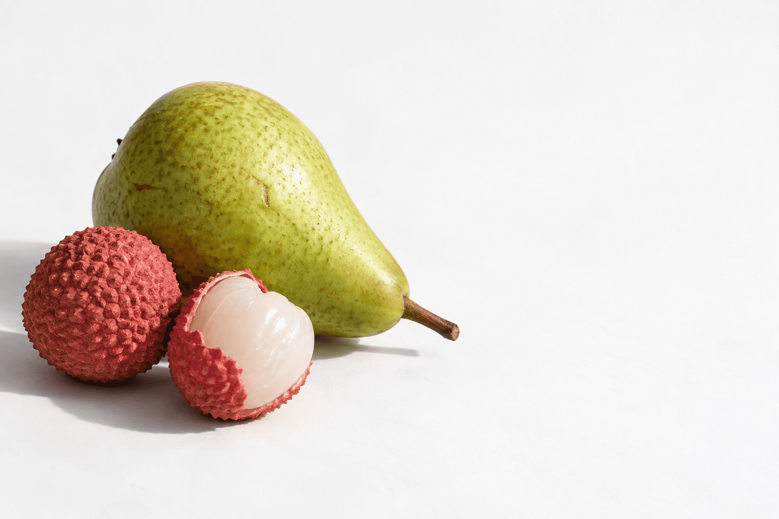A green pear resting on its side beside a whole lychee and a partly peeled lychee showing translucent white flesh.