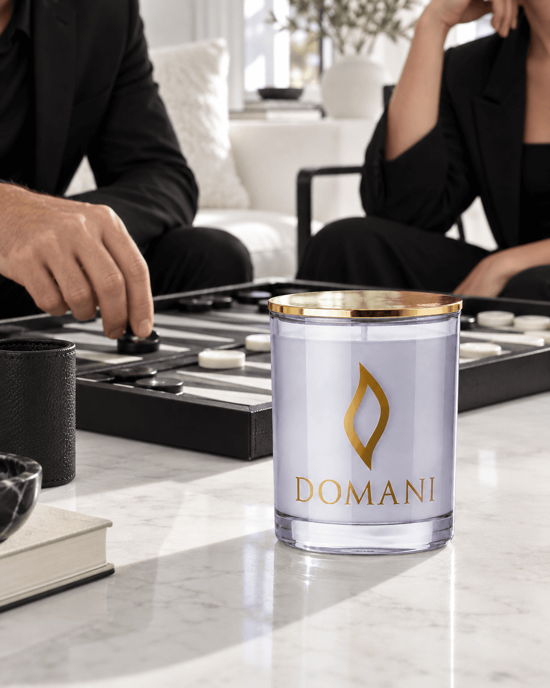 The unlit, lidded Domani candle in pale lavender Sapphire glass on a white game table, with two adults playing black-and-white backgammon behind it.