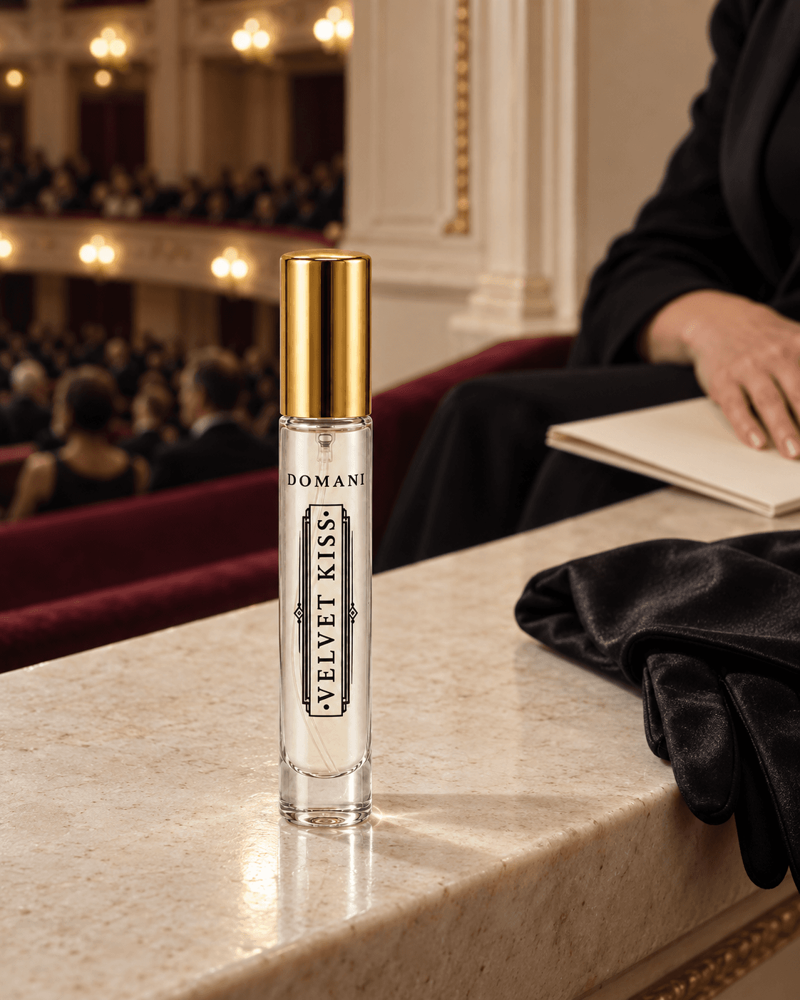 The capped Velvet Kiss 10 ml perfume on a pale stone concert-hall ledge beside a black satin glove, with a hand on a cream program and warm auditorium lights behind it.