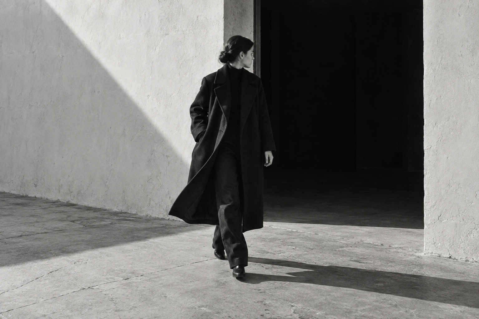 A woman in a long black coat steps across a pale concrete threshold beside a dark doorway, casting a sharp shadow across the floor.
